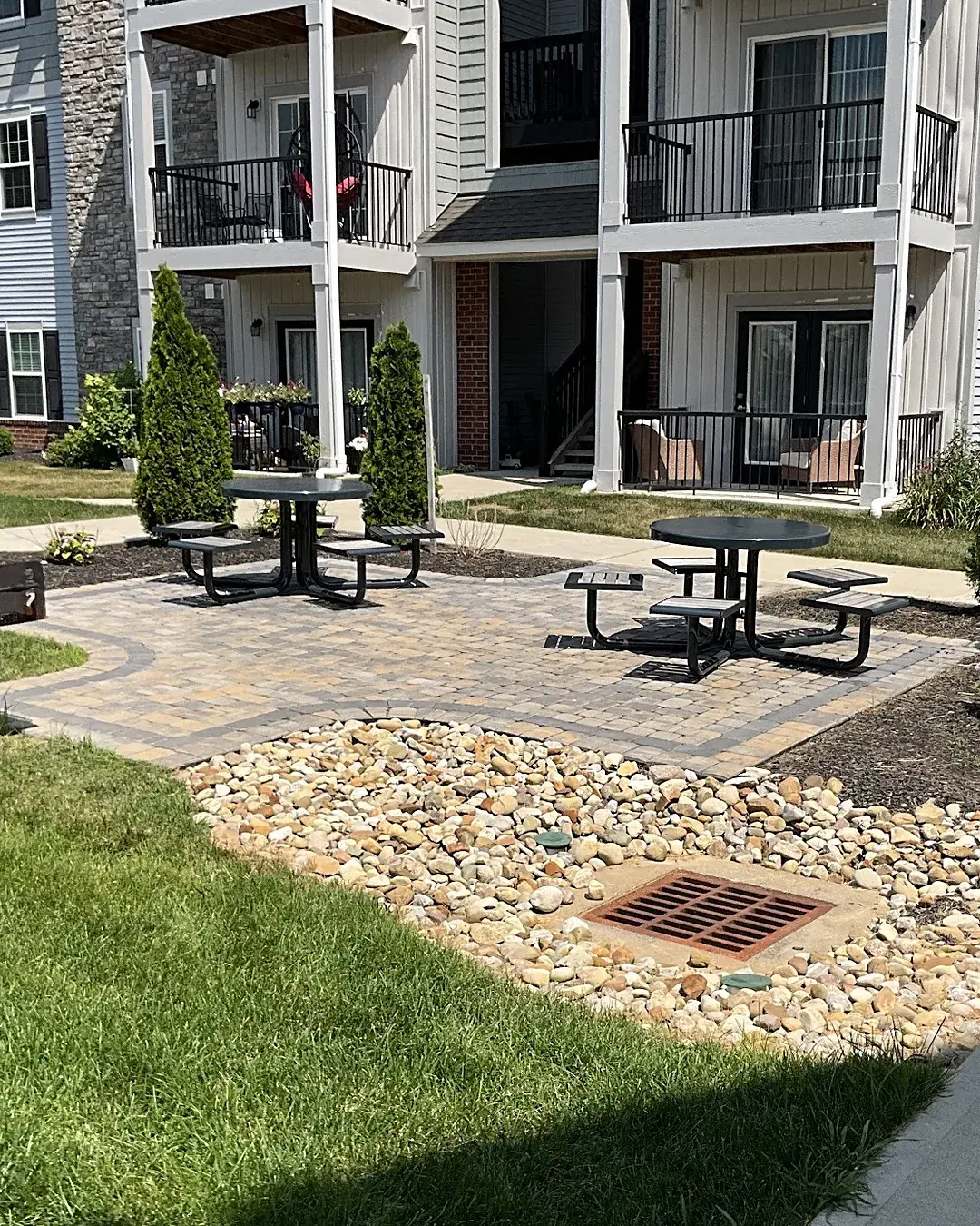 Apartment community courtyard with a paver patio, picnic tables, evergreen arborvitae and a river rock drainage bed