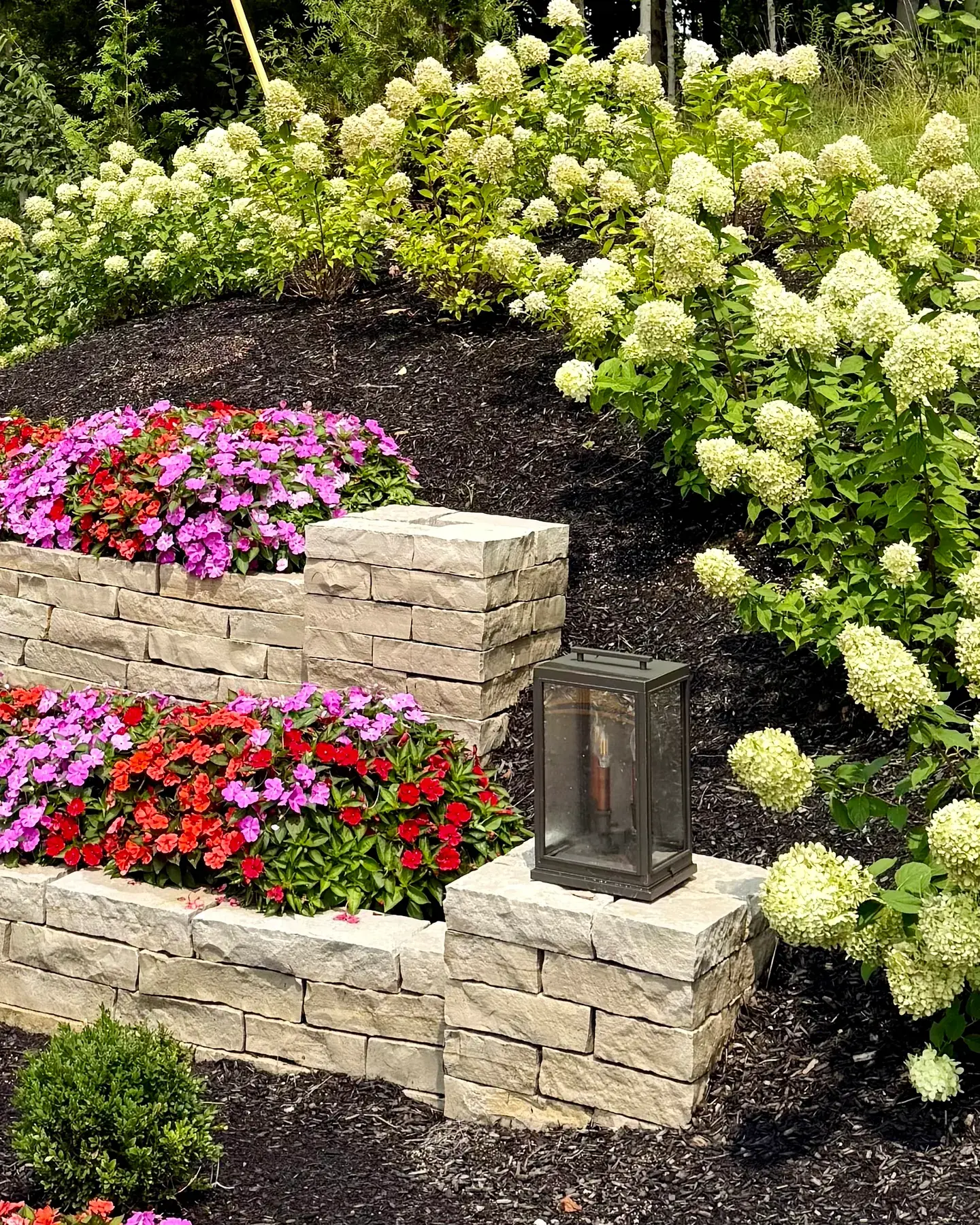 Tiered cut stone entrance walls planted with red, pink and purple impatiens, a lantern on the corner pier and limelight hydrangeas in full bloom at an apartment community