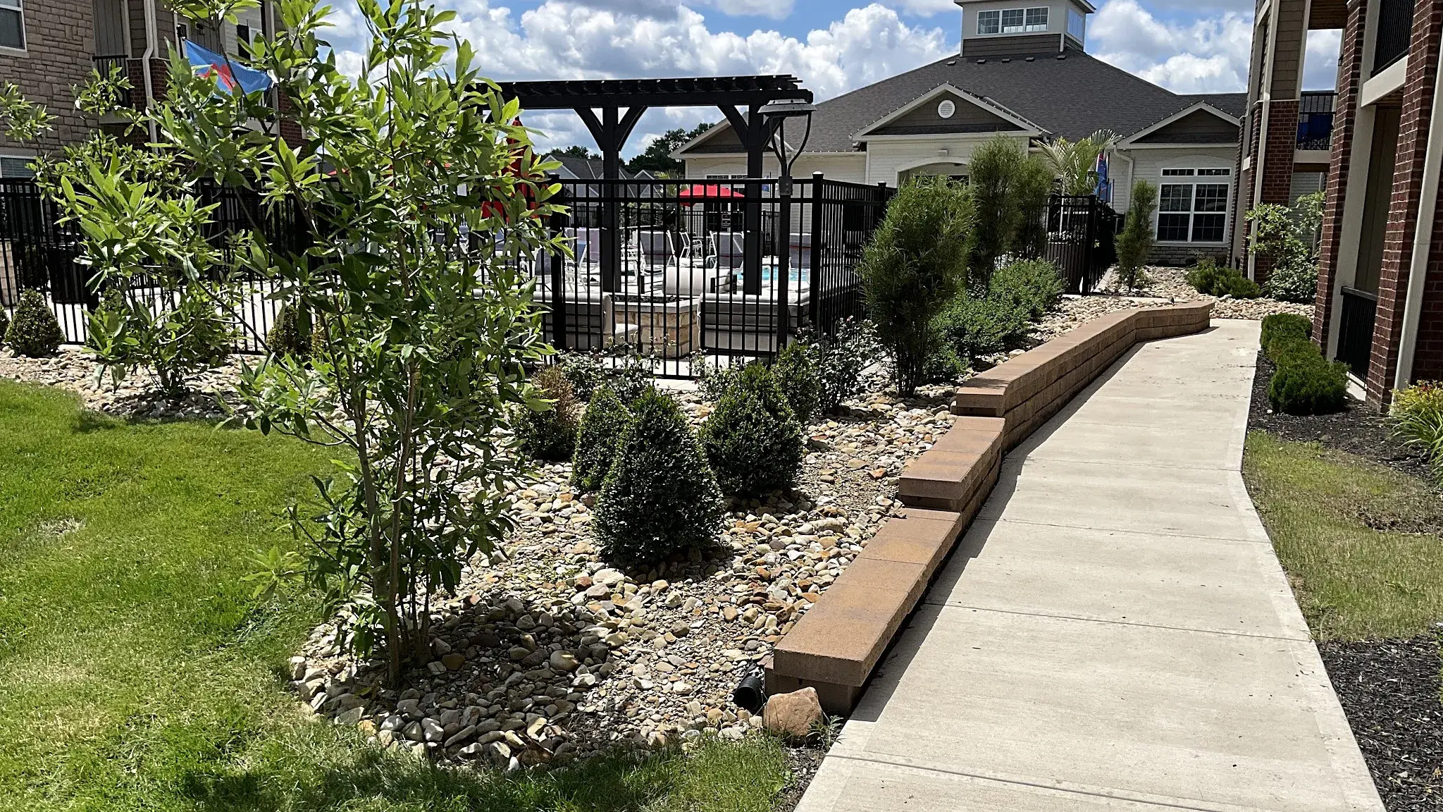 Apartment community pool courtyard with a river rock bed, clipped boxwoods, a young tree and a block wall beside a wide walkway