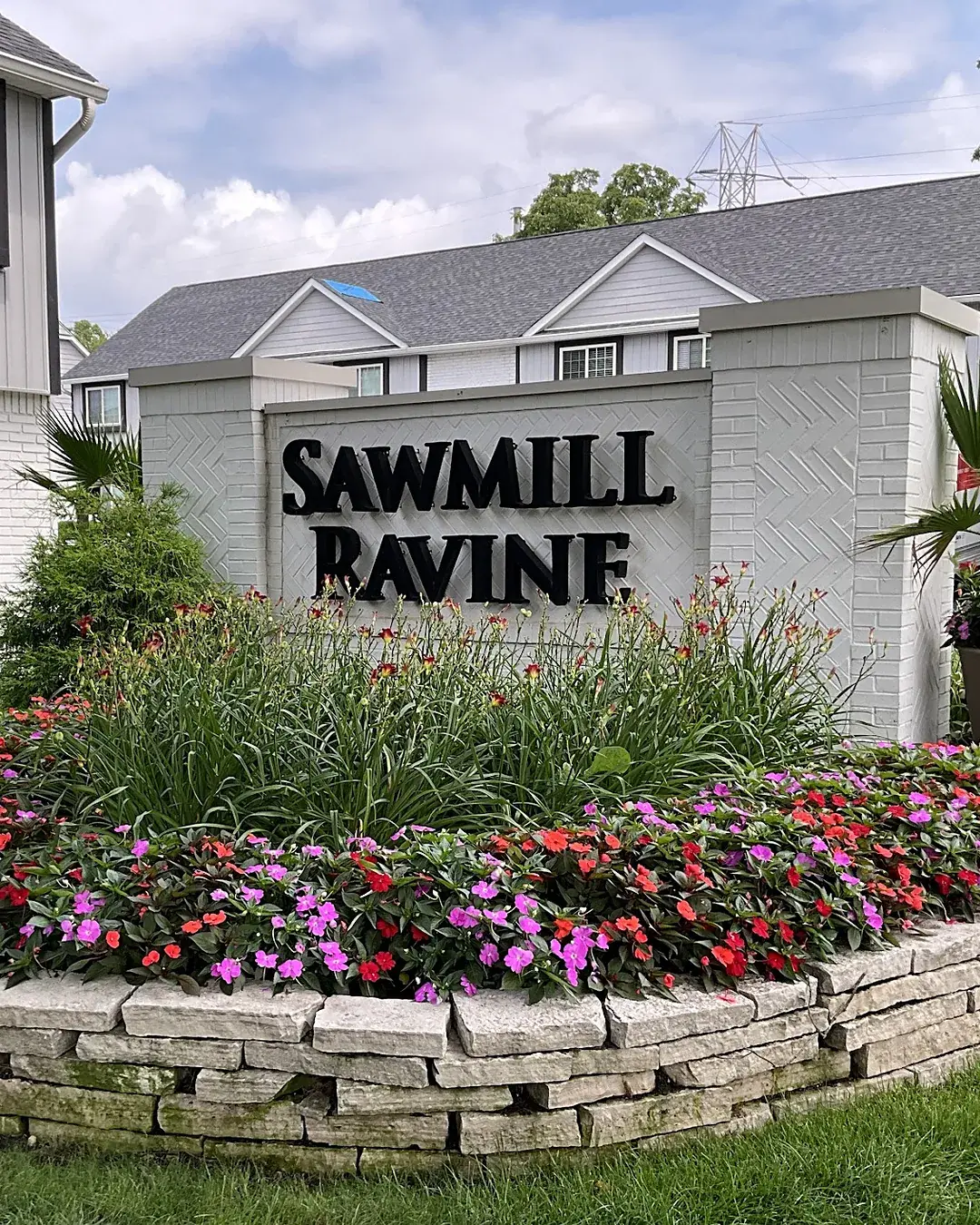 Apartment community entrance sign on a white brick wall in a stone-edged bed of impatiens, daylilies and a potted palm