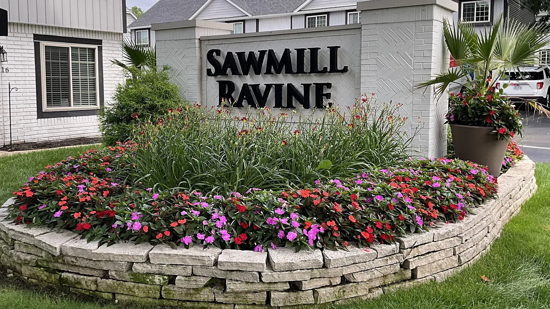 Apartment community entrance sign on a white brick wall in a stone-edged bed of impatiens, daylilies and a potted palm