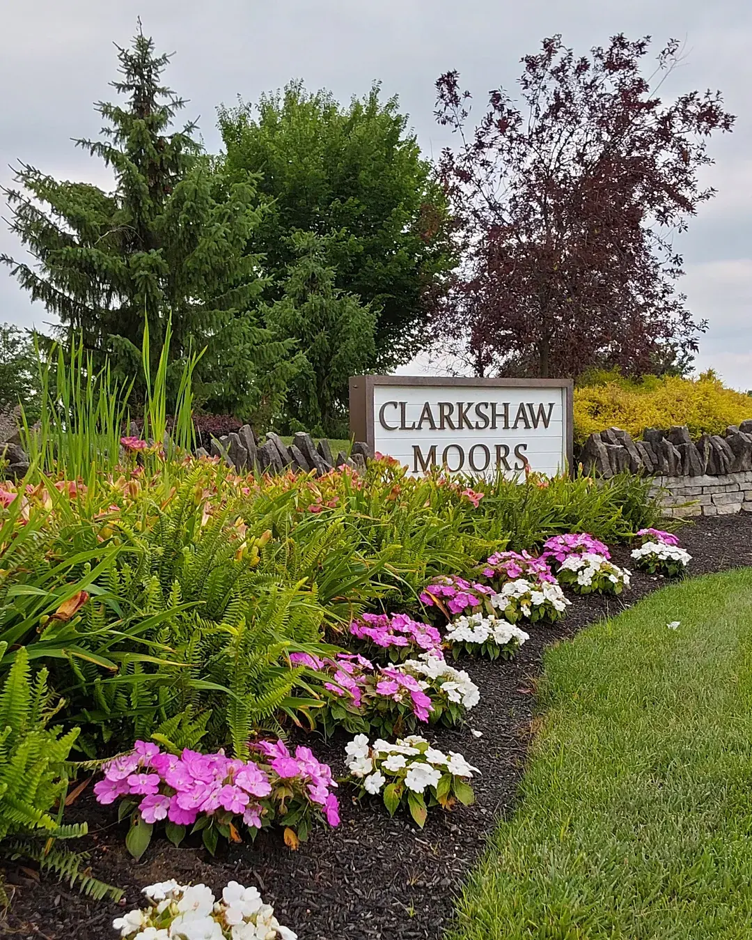 Curving mulched bed of ferns, daylilies and pink and white impatiens leading to an HOA entrance sign beside a mowed lawn
