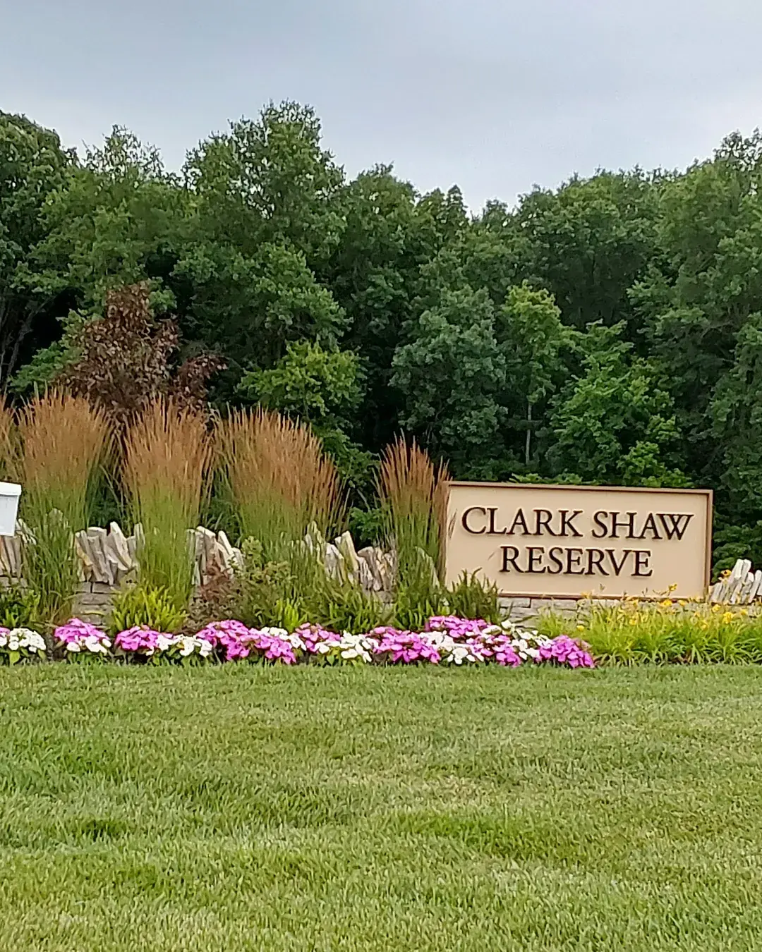 HOA community entrance with a stone pillar, wood sign, ornamental grasses and a row of pink and white annuals along a stacked stone wall