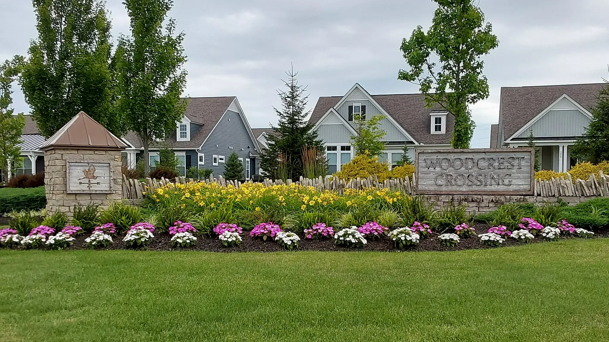 HOA community entrance with a stone pillar, rustic wood sign and a long bed of yellow daylilies and pink and white annuals