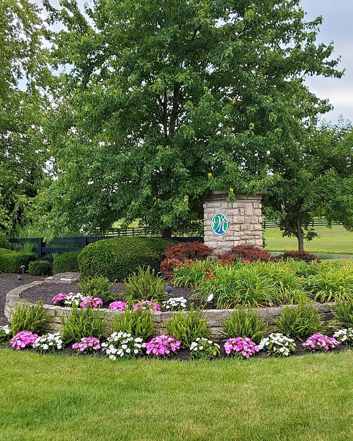 Round stone-edged entrance bed of impatiens, ferns and daylilies around a stone monument, under mature shade trees at a residential community