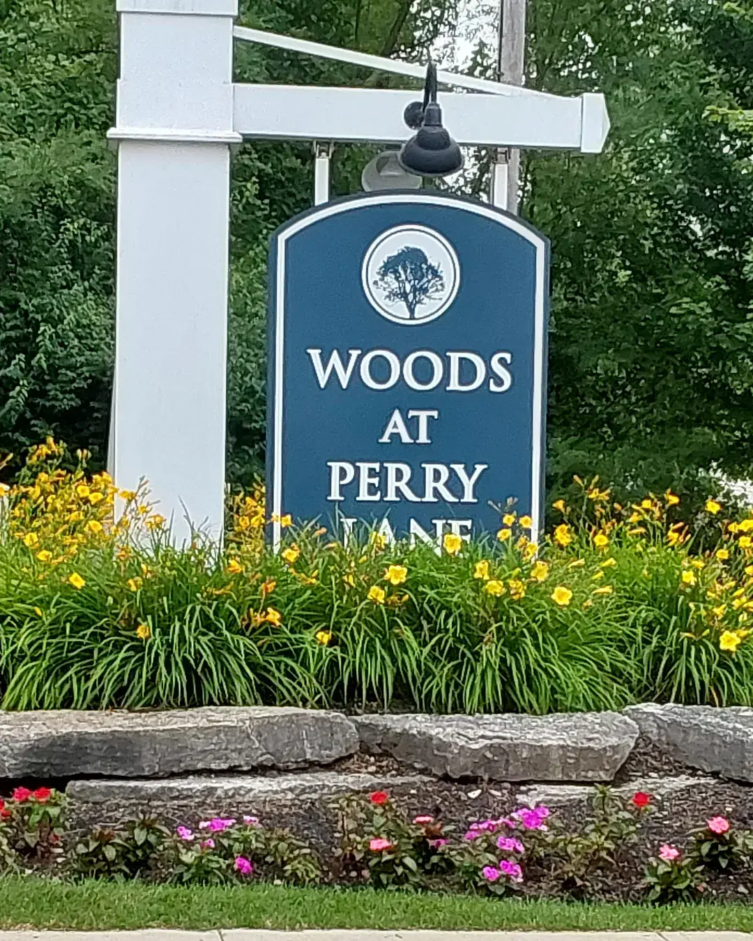 HOA community entrance sign on a white post above a tiered natural stone wall with blooming daylilies and impatiens