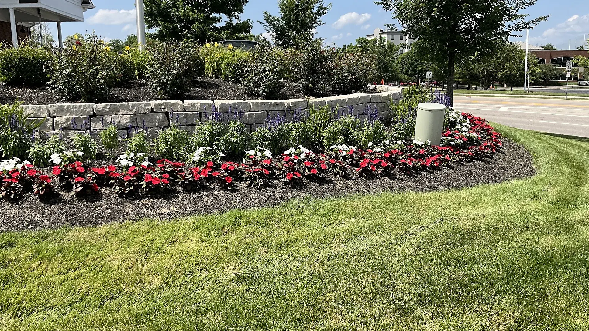 Landscaped island at a commercial property with a stone retaining wall, mixed perennials and a border of red and white begonias over fresh mulch