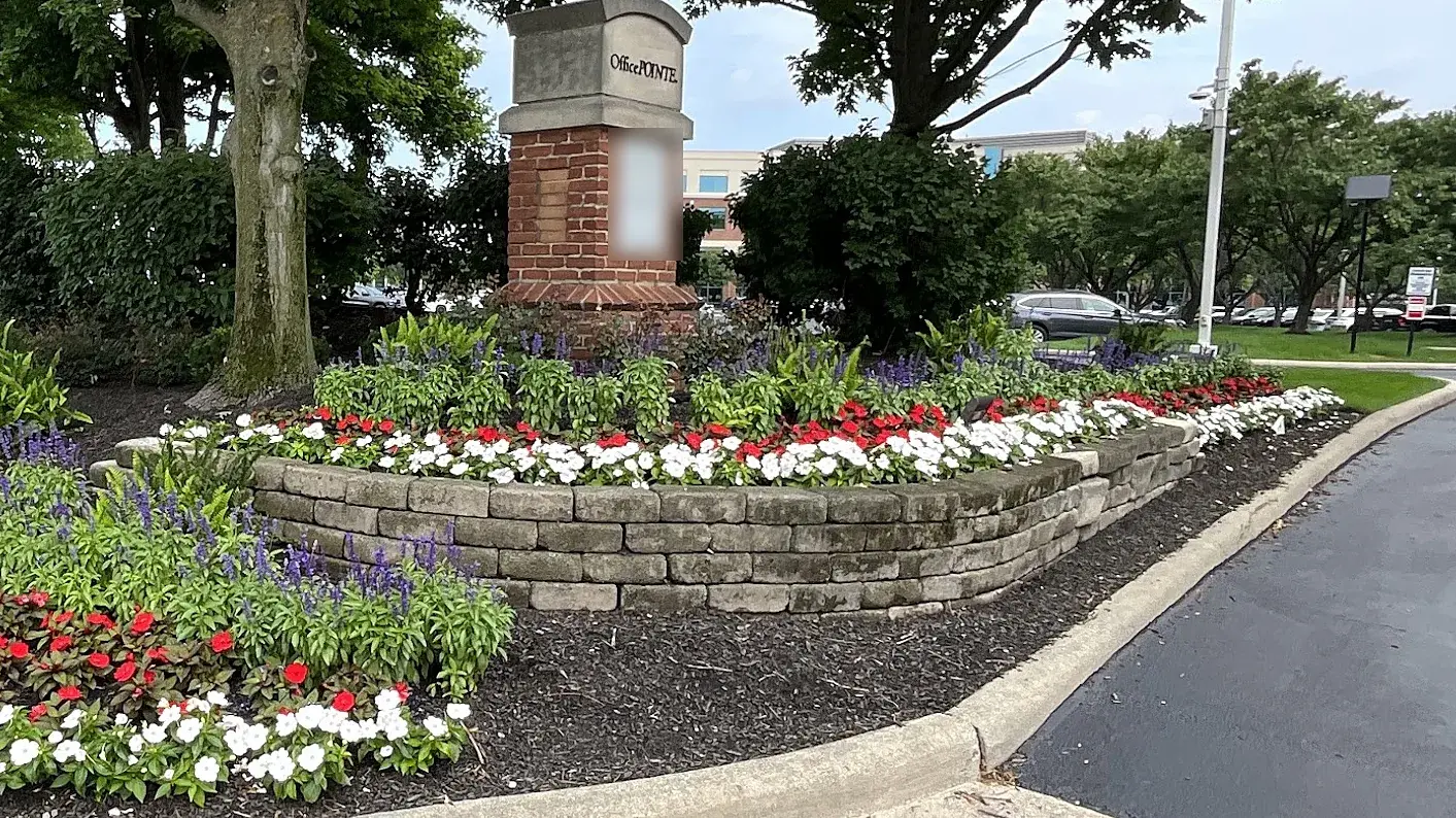 Office park entrance monument with a curved stone-edged bed of white and red begonias and blue salvia beneath mature shade trees