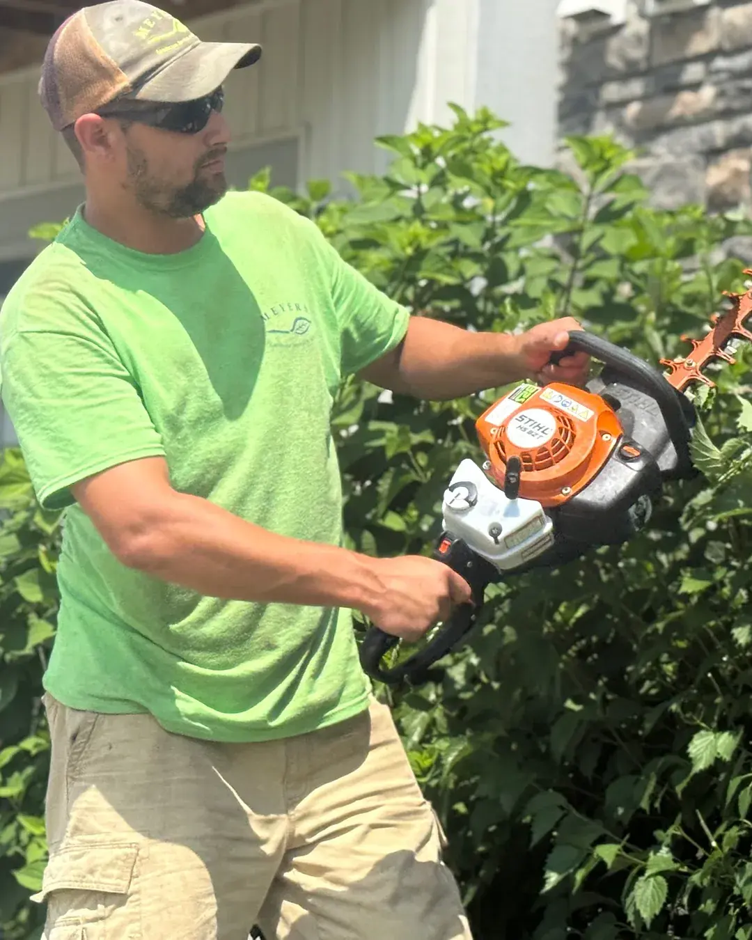Meyers crew member in a branded shirt and cap trimming a hedge with a power hedge trimmer
