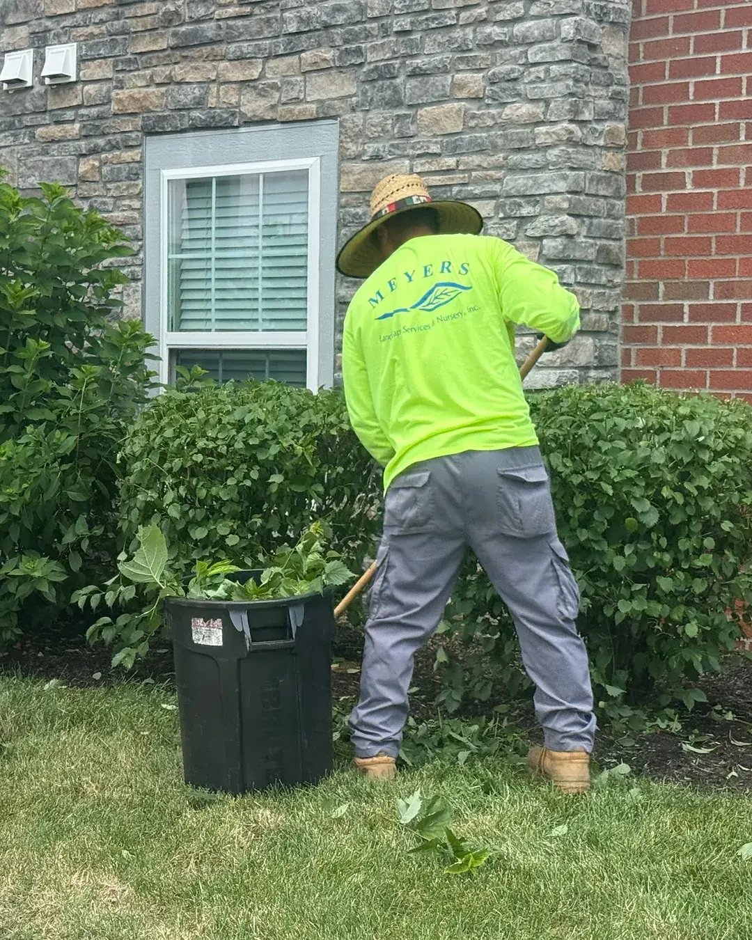 Two Meyers crew members in branded shirts and hats pruning shrub beds along the wall of an apartment building