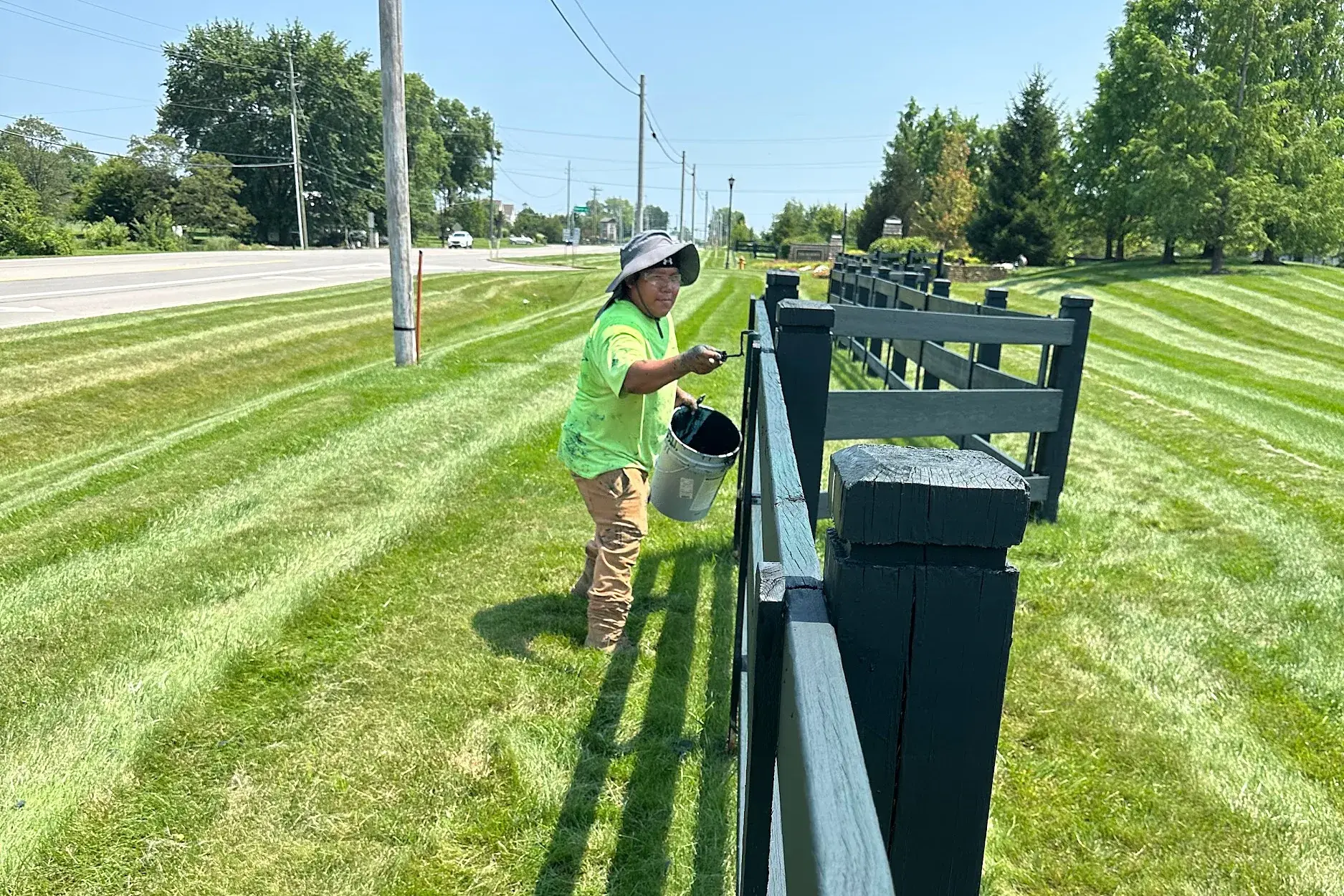 Crew member working along a rail fence beside a freshly striped lawn at a roadside community frontage