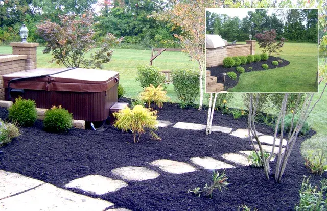 Backyard hot tub surrounded by black mulch beds, stepping stones, and young trees
