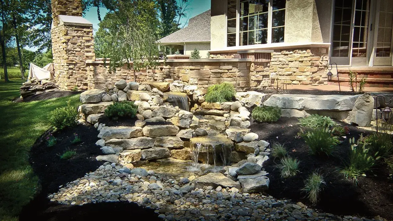 Tiered stone waterfall spilling into a pebble-lined basin beside a stone-walled patio