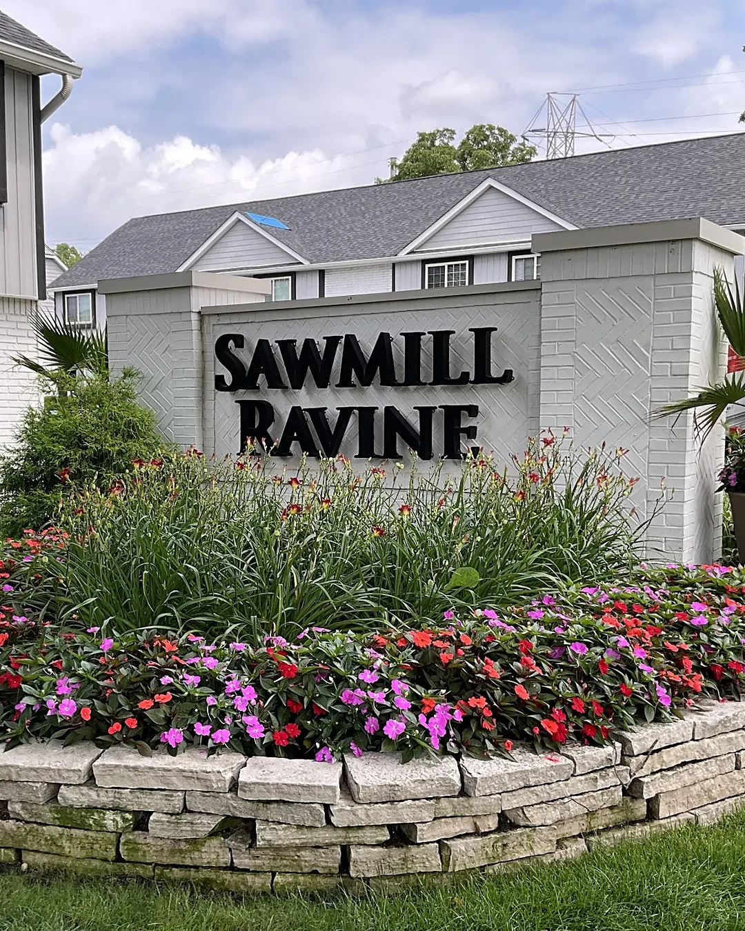 Apartment community entrance sign on a white brick wall in a stone-edged bed of impatiens, daylilies and a potted palm