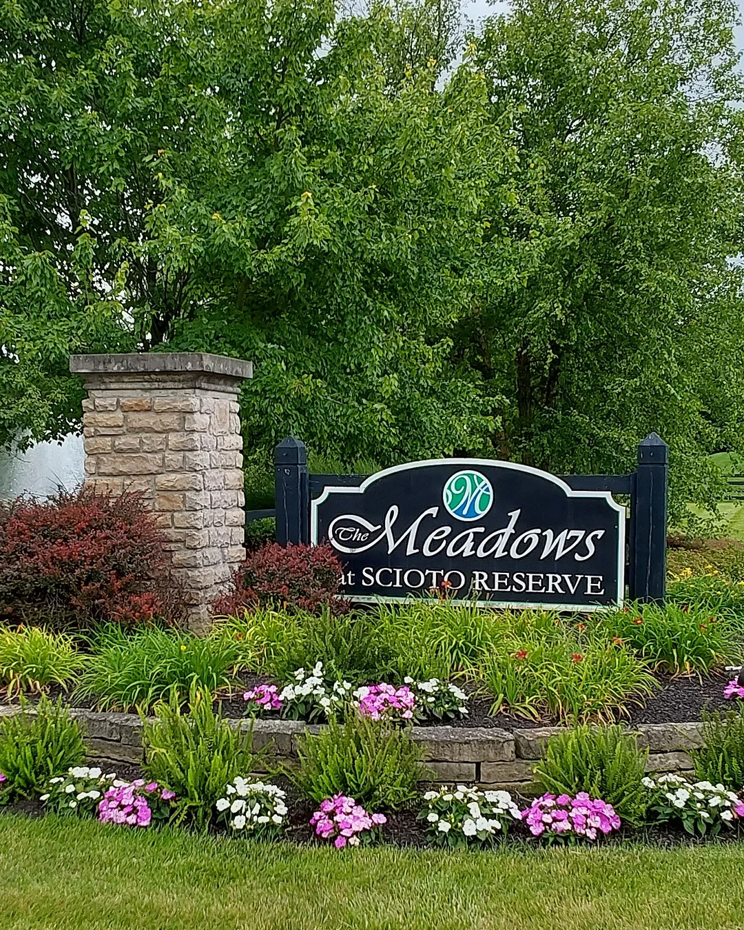 HOA community entrance monument with a navy sign, stone pillar and a round stone-edged bed of daylilies, grasses and pink and white impatiens beside a striped lawn