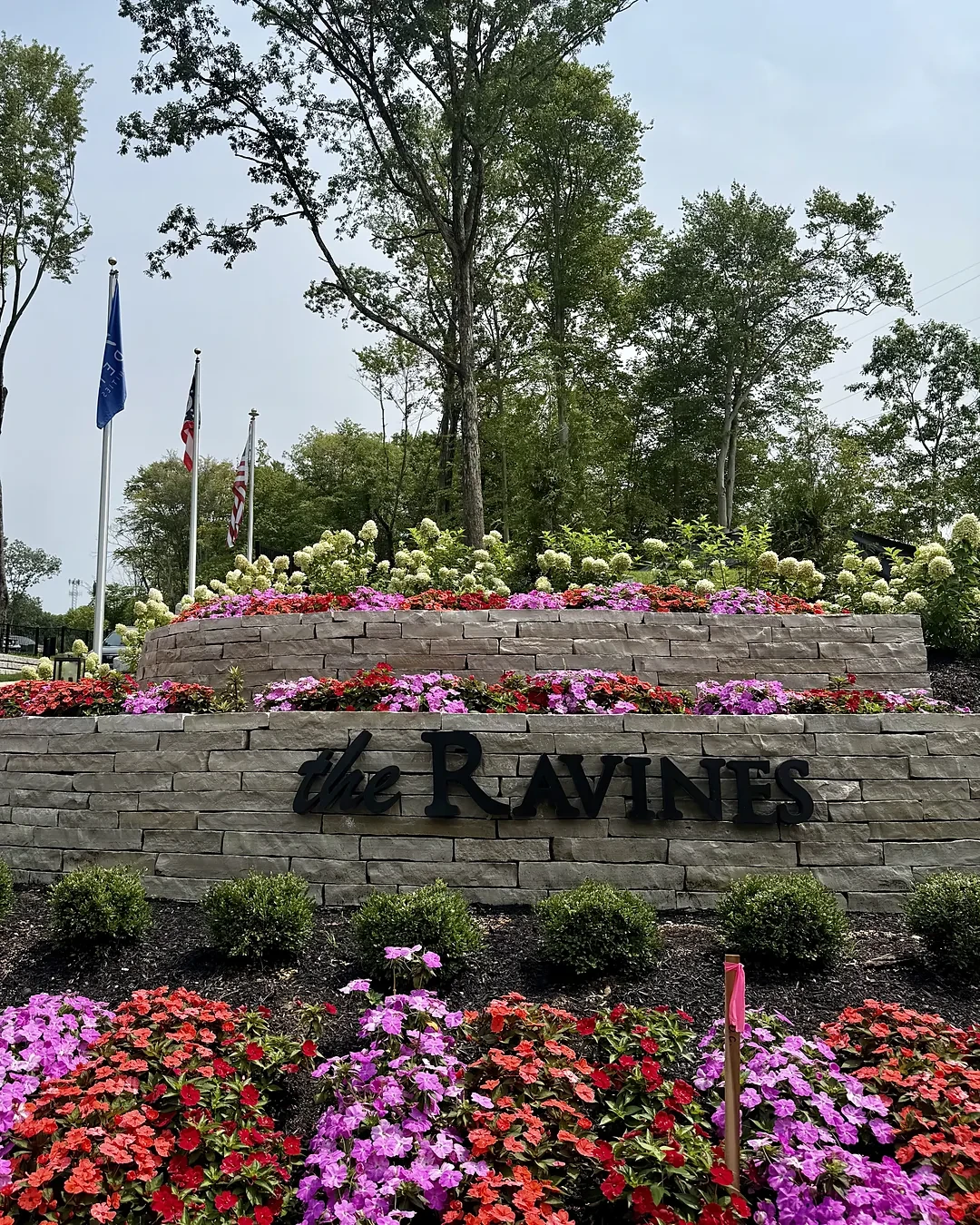Tiered natural stone entrance monument at an apartment community with flags, white hydrangeas and bright red, pink and purple impatiens