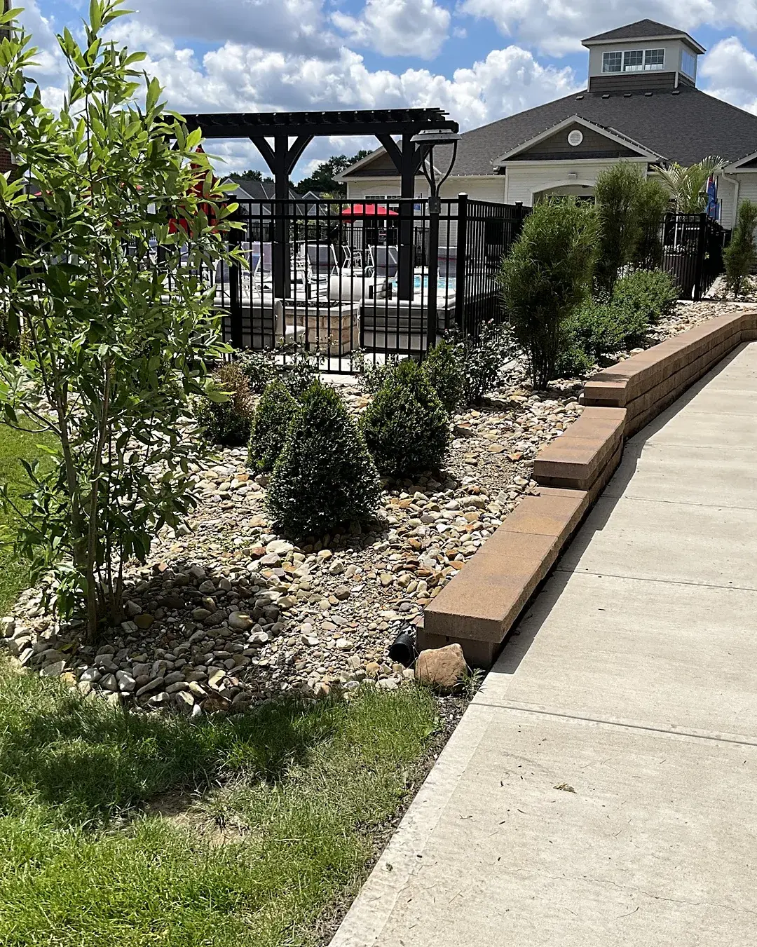 Apartment community pool courtyard with a river rock bed, clipped boxwoods, a young tree and a block wall beside a wide walkway