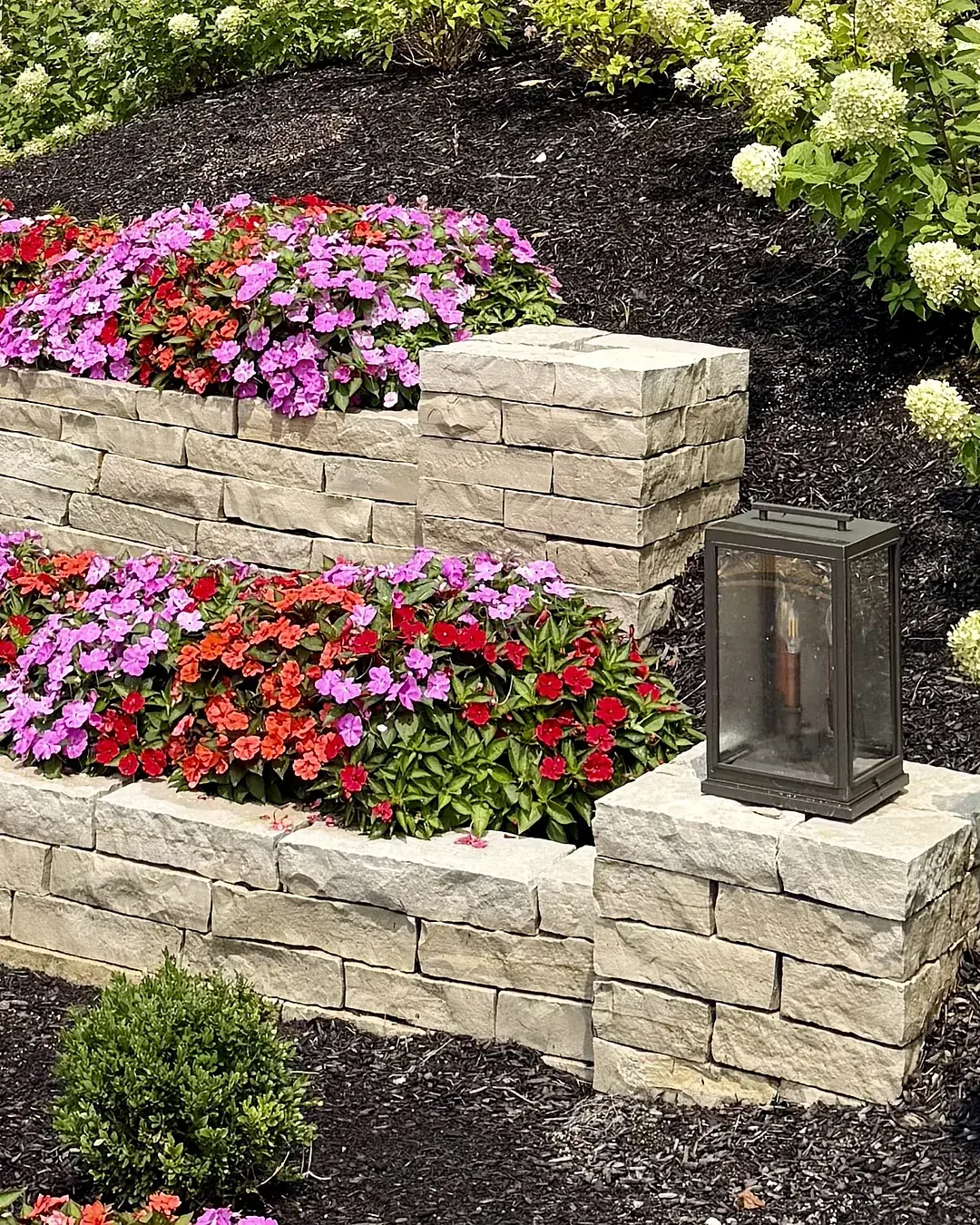 Two-tier natural stone wall with red, pink and purple impatiens and blooming white hydrangeas beside a lantern pier at a commercial property entrance