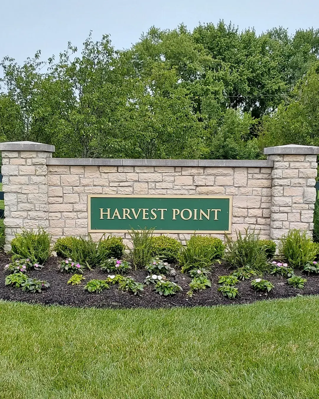 Stone entrance monument with a green sign above a crisp-edged mulch bed of ferns and young impatiens and clipped boxwoods