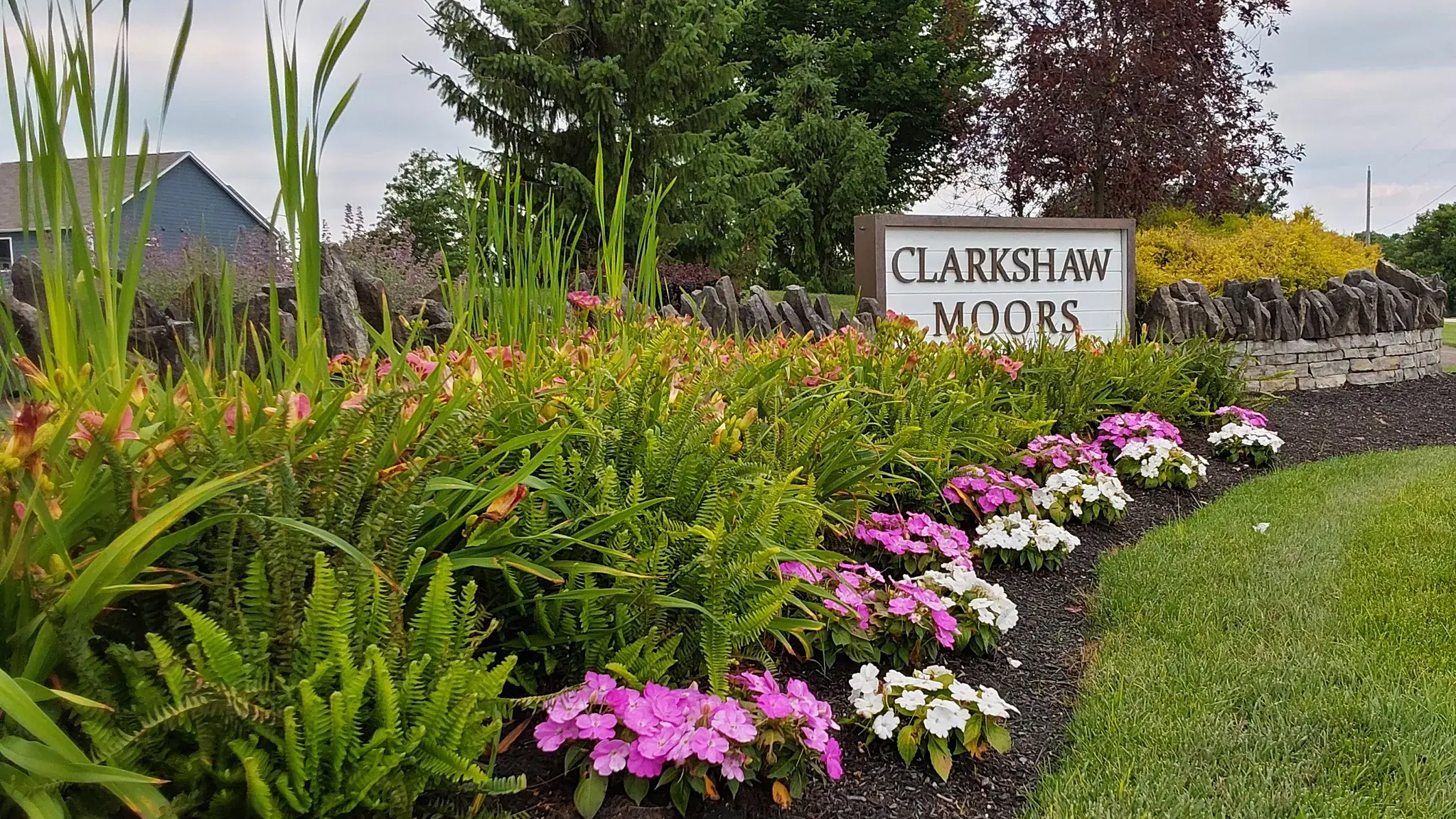 Curving mulched bed of ferns, daylilies and pink and white impatiens leading to an HOA entrance sign beside a mowed lawn