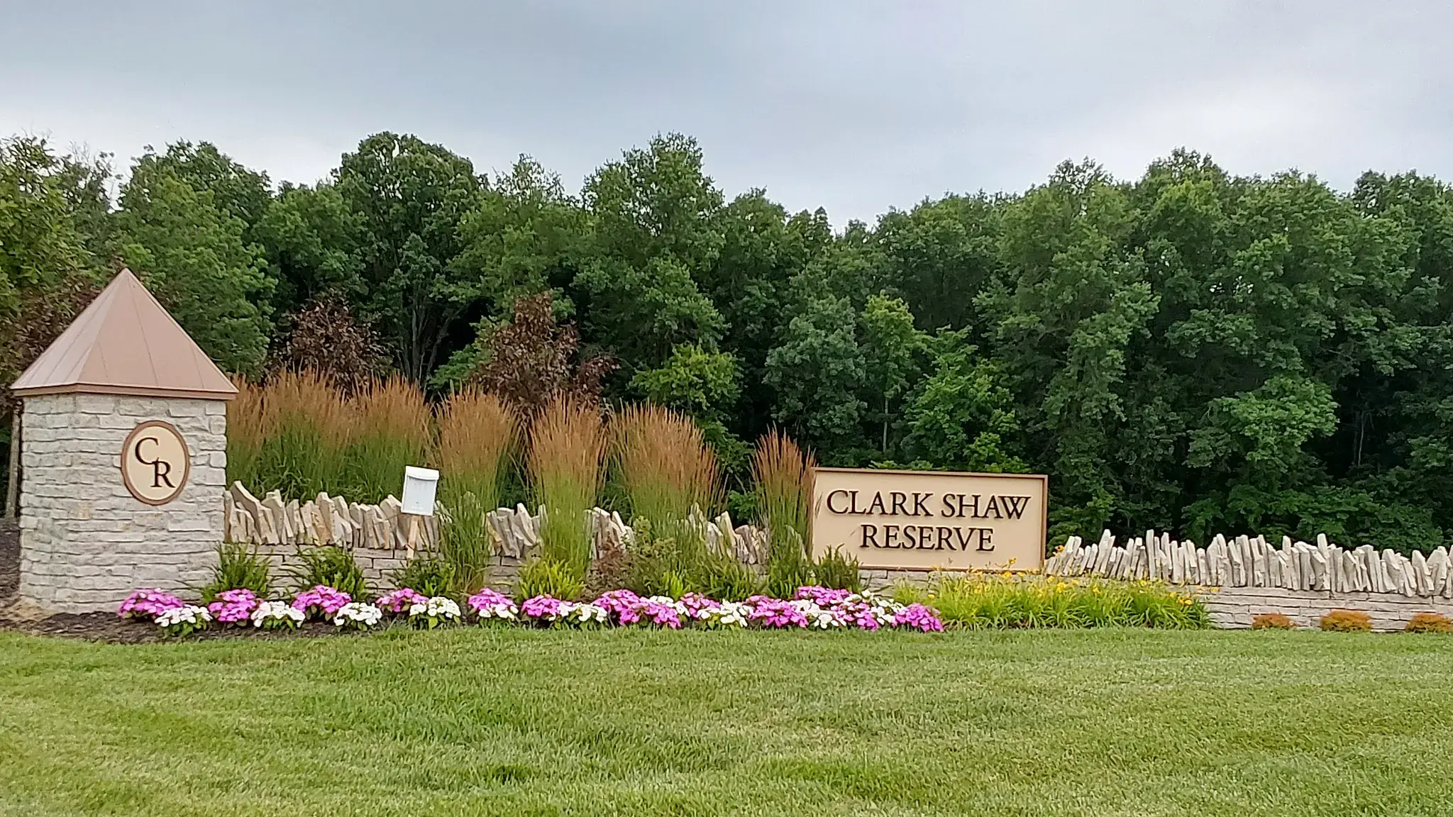 HOA community entrance with a stone pillar, wood sign, ornamental grasses and a row of pink and white annuals along a stacked stone wall