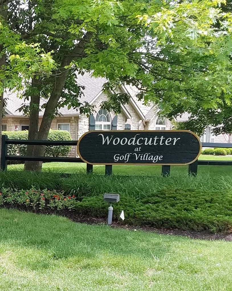 HOA community entrance with a stone pillar, rail fence sign, clipped hedge and a mulched annual bed under mature trees