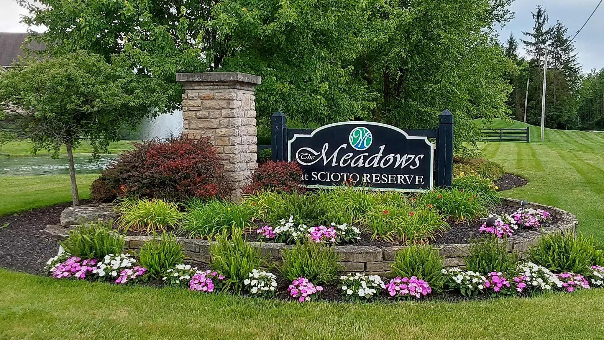 HOA community entrance monument with a navy sign, stone pillar and a round stone-edged bed of daylilies, grasses and pink and white impatiens beside a striped lawn