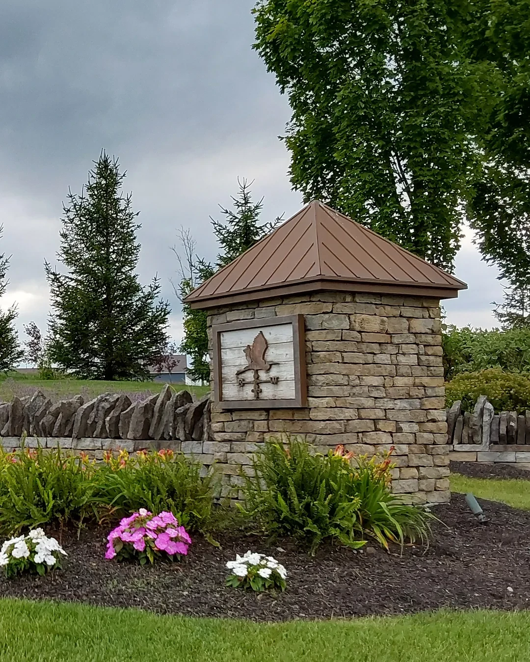 HOA community entrance with a stone sign pillar, stacked stone wall, evergreens and a bed of pink and white annuals and daylilies
