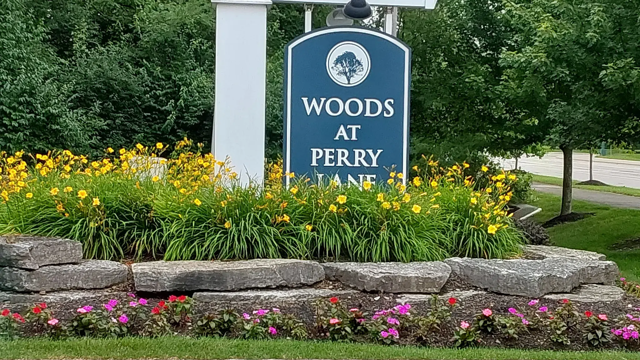 HOA community entrance sign on a white post above a tiered natural stone wall with blooming daylilies and impatiens