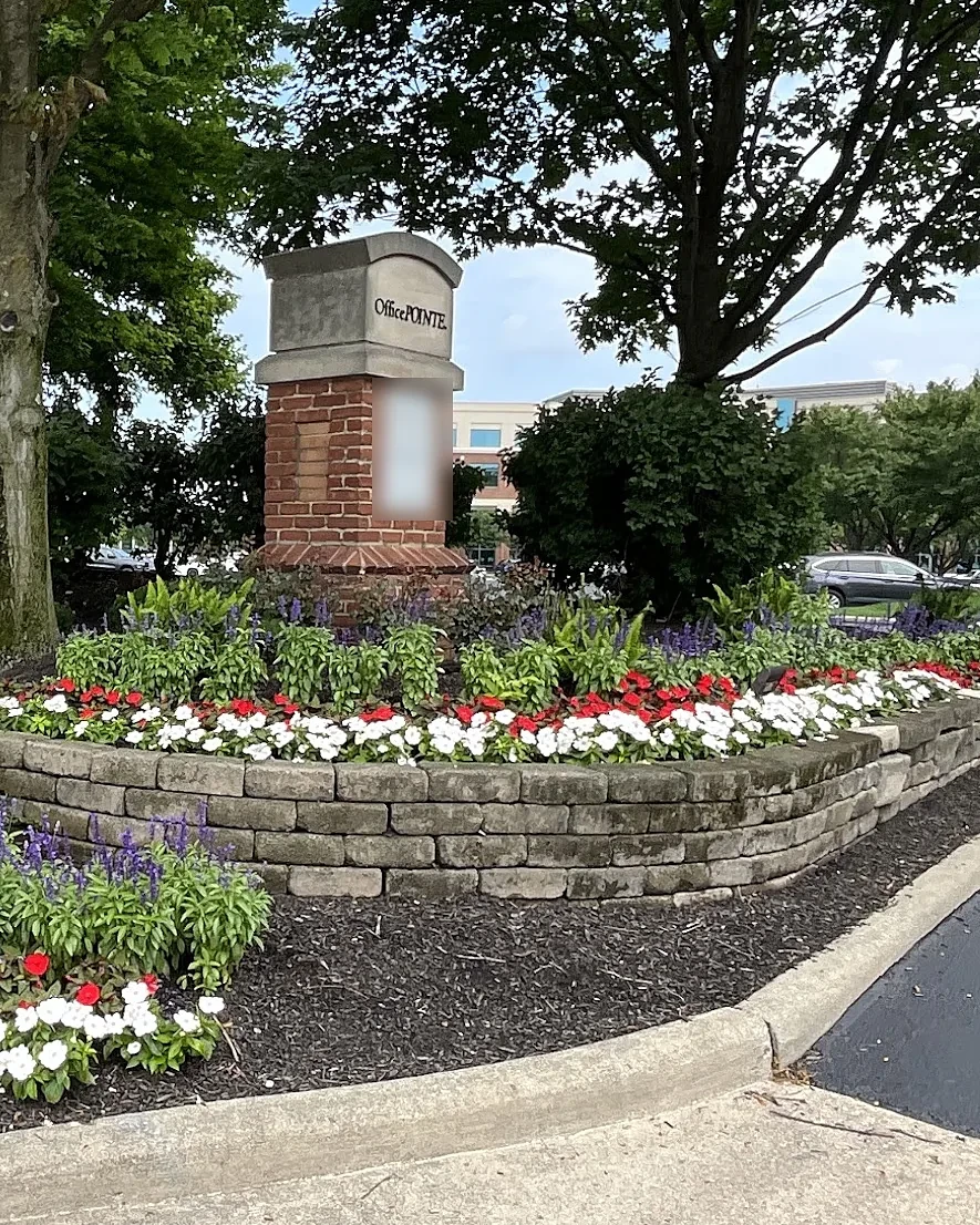 Office park entrance monument with a curved stone-edged bed of white and red begonias and blue salvia beneath mature shade trees