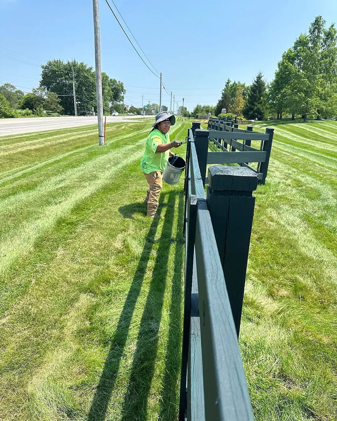 Crew member working along a rail fence beside a freshly striped lawn at a roadside community frontage